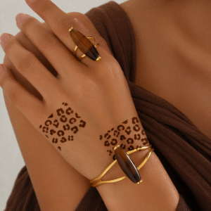 Close-up of hands with leopard print henna designs on the back of the hand and wrist, styled with gold rings and bracelets against a warm brown background.