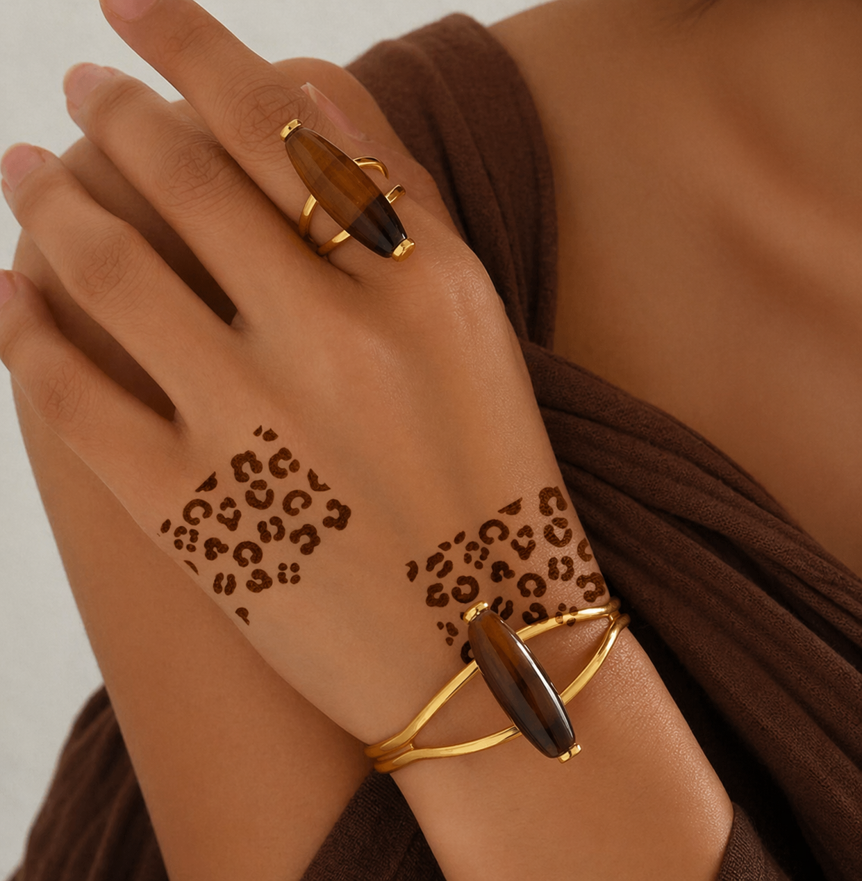 Close-up of hands with leopard print henna designs on the back of the hand and wrist, styled with gold rings and bracelets against a warm brown background.