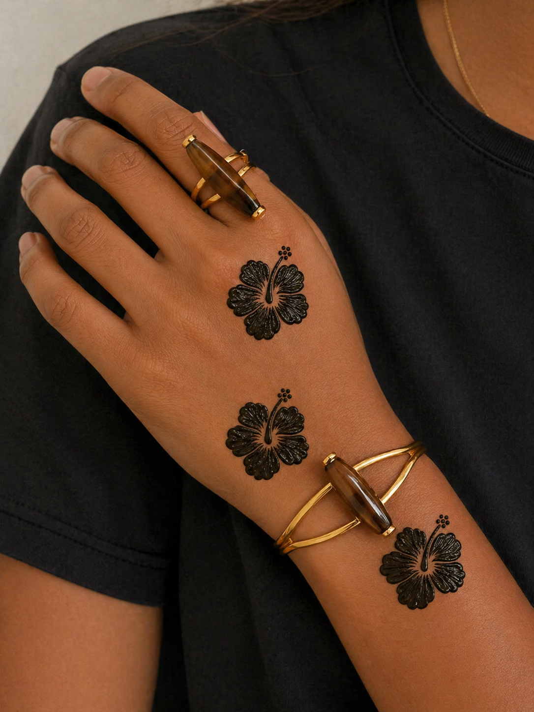 Woman wearing black clothing with Hibiscus floral henna designs on the back of her hand and forearm, paired with gold rings and bracelets.